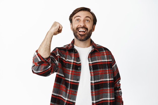 Joyful bearded man watching game and cheering rooting for team raising clenched fist and looking up happy standing over white background