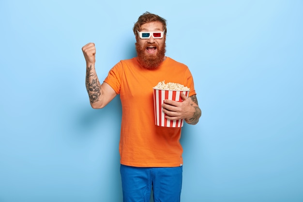 Радостный бородатый рыжий мужчина ест попкорн