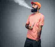 Free photo joyful bearded tattooed male smokes electronic cigarette isolated on grey background.