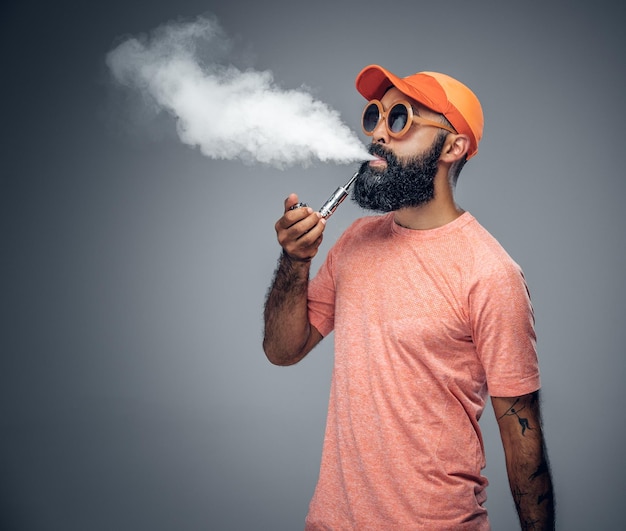 Радостный бородатый татуированный мужчина курит электронную сигарету, изолированную на сером фоне.