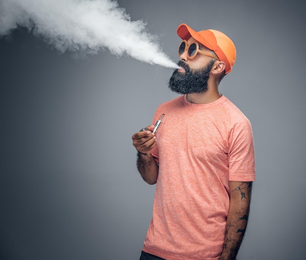 Joyful bearded tattooed male smokes electronic cigarette isolated on grey background.