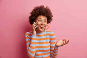 Бесплатное фото Радостная красивая афроамериканка разговаривает по телефону, наслаждается разговором, смотрит в сторону с милой улыбкой, держит руку поднятой, носит повседневный полосатый джемпер, изолированный над розовой стеной, имеет интересный разговор