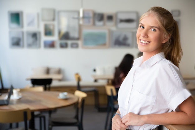 Joyful beautiful blonde woman wearing white shirt, standing in co-working space, leaning on desk, posing, 