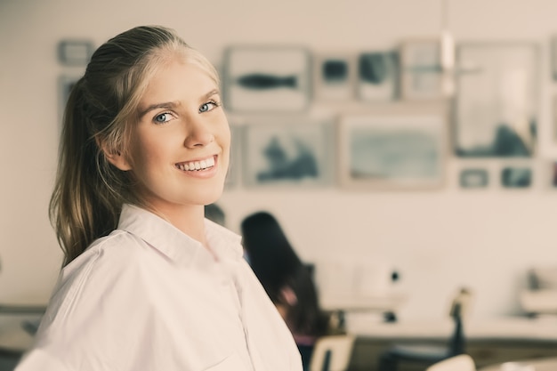 Free photo joyful beautiful blonde woman wearing white shirt, standing in co-working space, leaning on desk