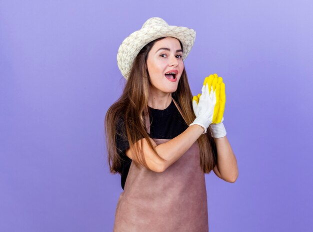 Joyful beautiful gardener girl in uniform wearing gardening hat and gloves showing pray gesture isolated on blue