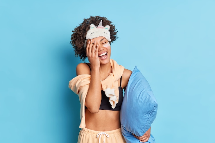Free Photo joyful beautiful woman curly hair covers face with palm feels very happy dressed in nightwear holds pillow under arm isolated over blue wall