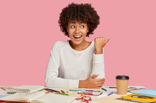 うれしそうな黒人の創造的な女性は前向きな表情をしていて、親指で脇を向いて、広告のための空きスペースを示し、ピンクの壁に隔離されたスパイラルノートとクレヨンで職場でポーズをとる