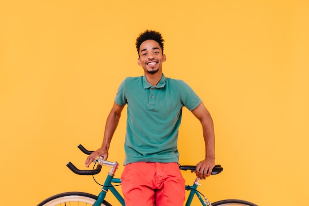 Радостный черный велосипедист смеется. Красивый африканский молодой человек с удовольствием позирует возле своего велосипеда.