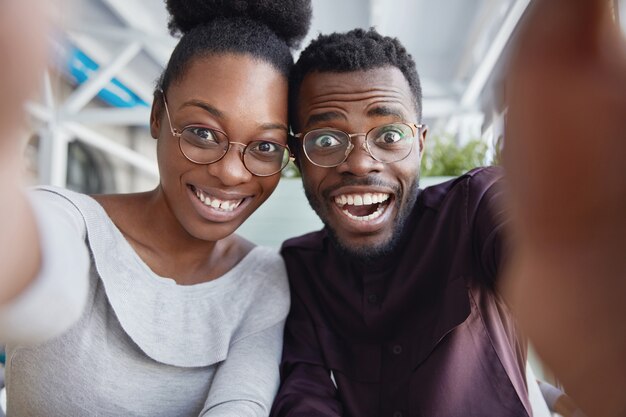 Joyful black male and female best friends have fun together, take picture of themselves or pose for making selfie, being in good mood after successful day.