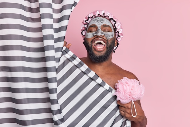 Joyful black man laughs while taking shower holds bath sponge enjoys grooming procedures poses behind curtain applies facial clay mask isolated on pink wall in bathroom