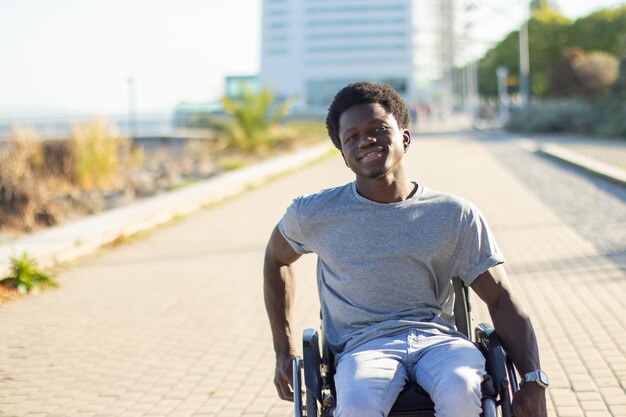 Joyful Black man in wheelchair enjoying warm summer day in city park while riding in park along seafront road. City building in background. Front view. Medium shot. Disability, attitude concept.