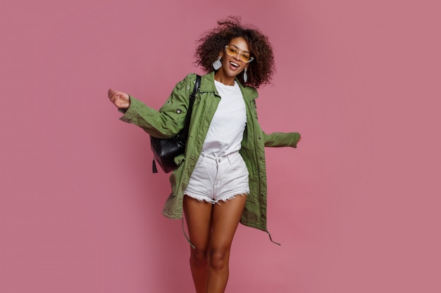 免费照片快乐的黑人妇女有乐趣在工作室在粉红色的背景。白色t恤，绿色夹克。时髦的春季装扮。