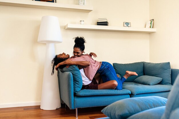 Joyful black young girlfriends hugging on couch at home