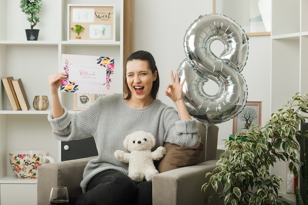 Joyful blinked showing okay gesture beautiful girl on happy women day holding greeting card sitting on armchair in living room