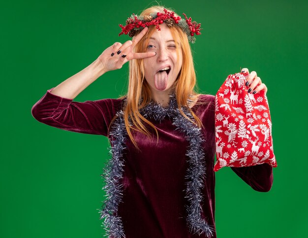 Радостная моргнула молодая красивая девушка в красном платье с венком и гирляндой на шее, держащая рождественскую сумку, показывая язык и делающая жест мира на зеленом фоне