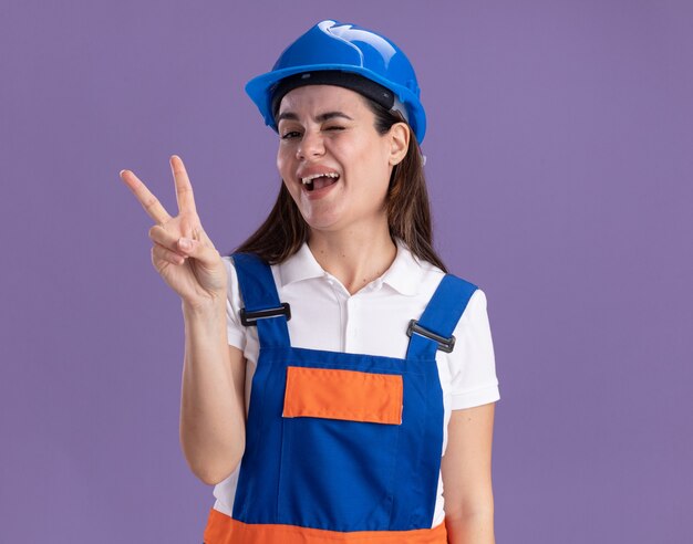 Радостный моргнул молодая женщина-строитель в униформе, показывающая жест мира на фиолетовой стене
