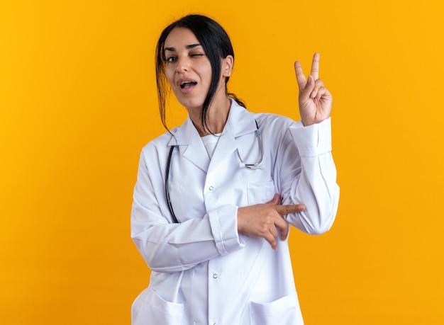 Радостная моргнула молодая женщина-врач в медицинском халате со стетоскопом, показывая жест мира, изолированный на желтой стене