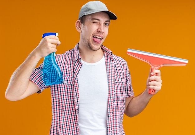 Joyful blinked young guy cleaner wearing cap holding cleaning agent with mop showing tongue head isolated on orange background