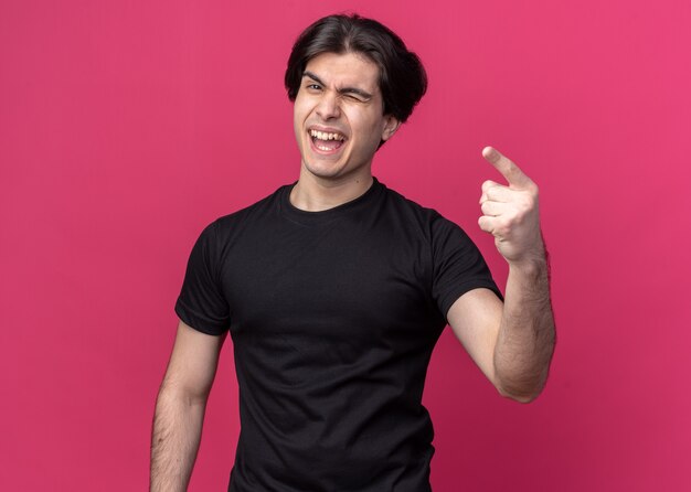 Joyful blinked young handsome guy wearing black t-shirt points isolated on pink wall