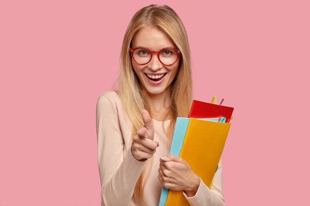 Joyful blonde female model wears round spectacles, makes gun gesture, holds textbooks
