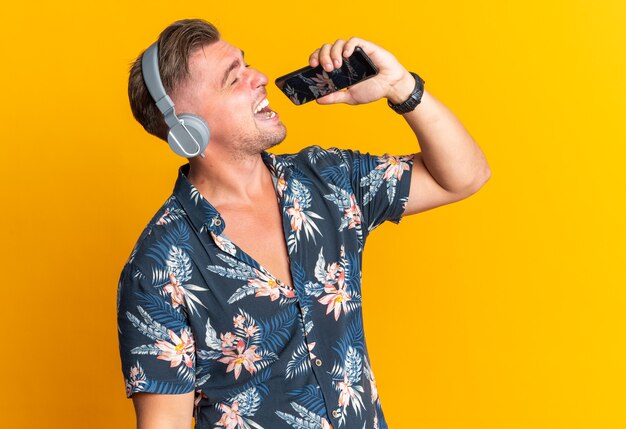 Joyful blonde handsome man on headphones holding phone close to his mouth pretending to sing isolated on orange wall with copy space