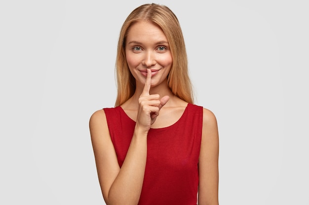 Free photo joyful blonde woman makes silence gesture, keeps fore finger on lips, asks to keep information confidential