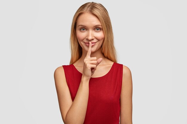 Joyful blonde woman makes silence gesture, keeps fore finger on lips, asks to keep information confidential
