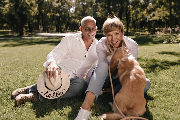 Joyful blonde woman in white blouse and jeans smiling, hugging dog and sitting with grey haired man with mustache and hat in shirt outdoor.