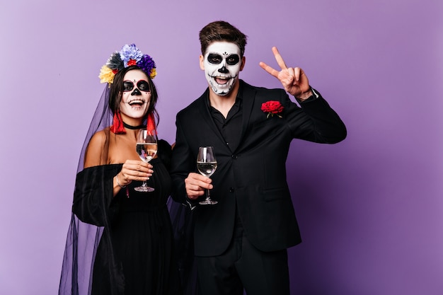 Joyful boyfriend and girlfriend drink champagne and celebrate halloween in image of mexican-style bride and groom