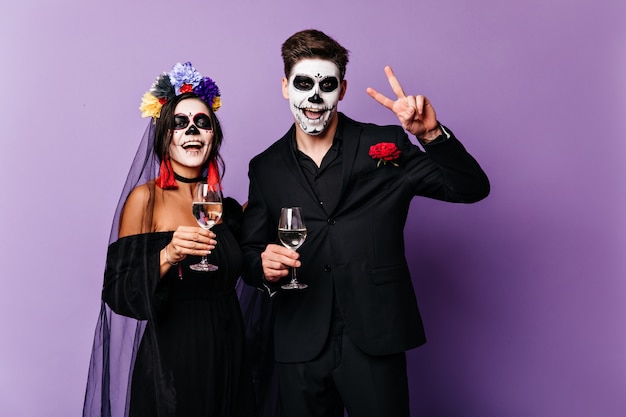Веселые парень и девушка пьют шампанское и празднуют Хэллоуин в образе жениха и невесты в мексиканском стиле.