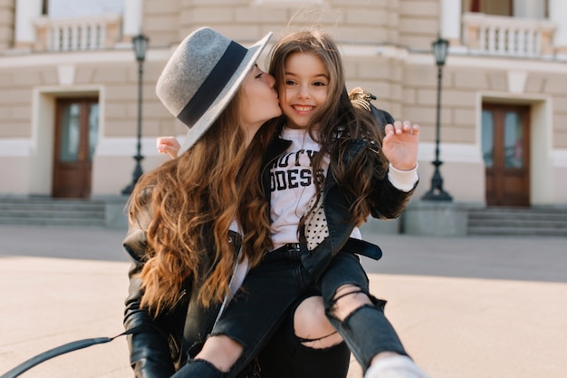 Joyful brunette girl with lovely face expression in stylish jeans with holes sitting on mom's knee and laughing. Beautiful woman wearing elegant hat kissing daughter in cheek in the middle of street.