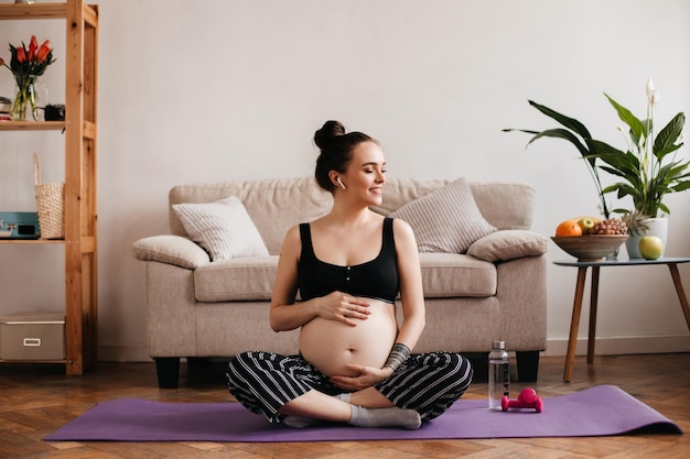 Joyful brunette pregnant woman in cropped top and black pants smiles listening to music in white wireless headphones Happy girl with tattoos meditating on purple carpet and gently touches belly