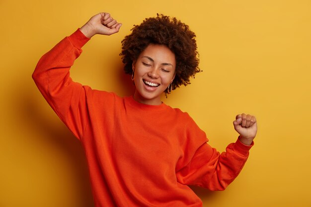 joyful brunette woman has fun and dances with hands raised, dressed in casual jumper, cheers over yellow background, gets promotion or approval, celebrates win.