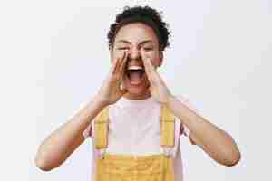 Free photo joyful carefree african american woman in yellow overalls, holding palms around opened mouth, shouting, calling someone on distance