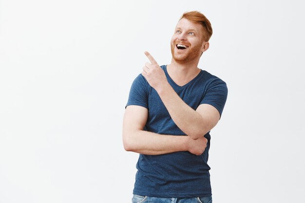 Joyful carefree and amused good-looking masculine redhead male with bristle in blue t-shirt, pointing and gazing at upper left corner