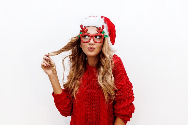 Joyful carefree blond woman in cute masquerade glasses and new year hat in red knitted sweater posing on white wall. Isolate. Christmas and new test party concept.