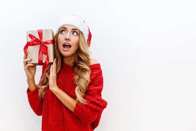 Joyful carefree blond woman in cute masquerade glasses and new year hat in red knitted sweater posing on white wall. Isolate. Christmas and new year party concept.