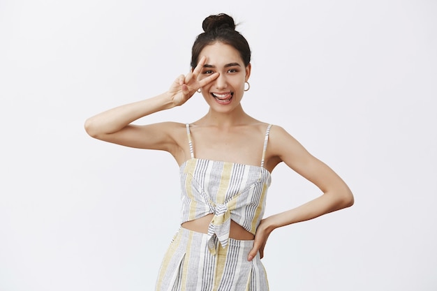 Joyful and carefree creative female designer in elegant outfit and bun hairstyle, showing peace or victory sign over eye, sticking out tongue playfully, holding hand on waist