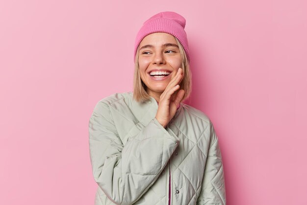 Joyful carefree teenage girl with happy expression smiles positively keeps hand on cheek being in good mood wears hat and jacket isolated over pink studio background Positive emotions concept