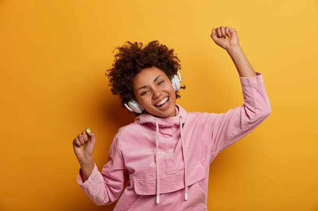 Joyful carefree woman dances to music, listens favourite audio track, raises hands with clenched fists, smiles broadly, wears rosy sweatshirt, isolated over yellow wall. People, leisure, entertainment