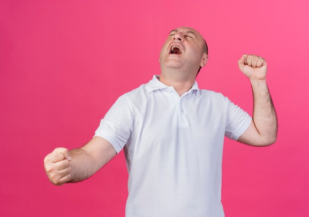 Joyful casual mature businessman clenching fists doing yes gesture with closed eyes isolated on pink background