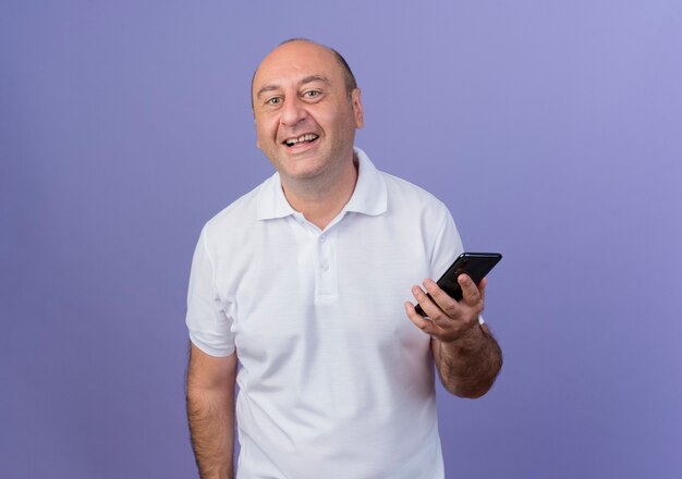 Joyful casual mature businessman holding mobile phone and looking at camera isolated on purple background with copy space