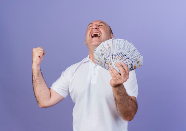 Joyful casual mature businessman holding money clenching fist doing yes gesture with closed eyes isolated on purple background with copy space