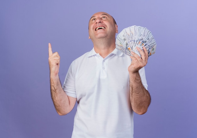 Joyful casual mature businessman holding money looking and pointing up isolated on purple background with copy space