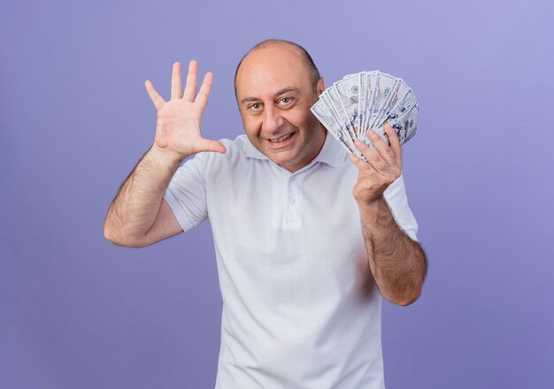 お金を保持し、コピースペースで紫色の背景に分離された手で5を示すうれしそうなカジュアル成熟したビジネスマン
