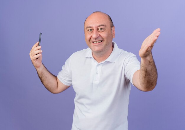 Gioioso casual imprenditore maturo guardando la telecamera tenendo il telefono cellulare e allungando la mano isolata su sfondo viola