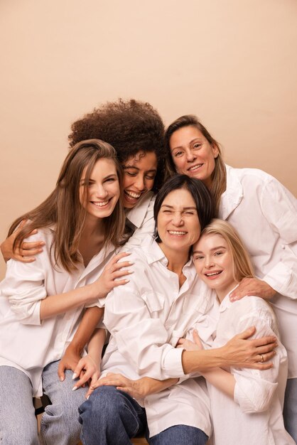 Joyful caucasian african women of different ages in white shirts laughing looking at camera on beige background