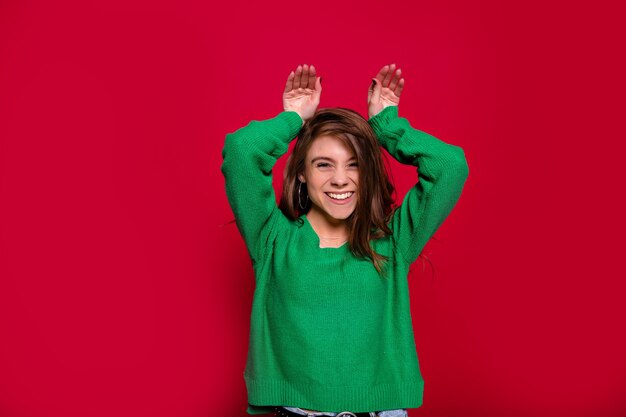 Joyful caucasian girl expressing positive emotions dressed green sweater while posing in new year on red background with holding up hands over head. Blithesome young woman in studio