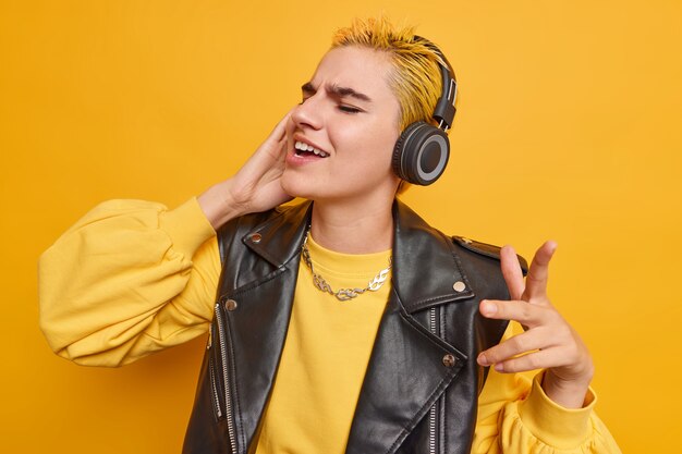 Joyful Caucasian girl with trendy hairstyle listens music in wirelesss headphones catches every bit of song dances to rhythm dressed in stylish clothes isolated over yellow wall