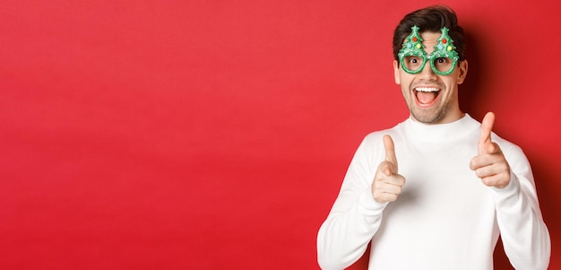 Joyful caucasian guy in party glasses and white sweater smiling and pointing fingers at camera wishing merry christmas and happy new year standing over red background